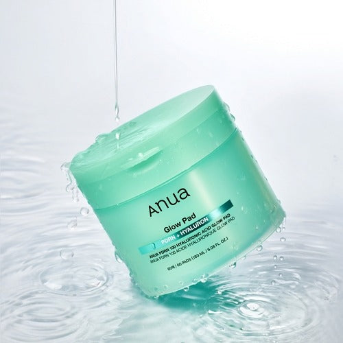 Green jar of Anua Glow Pad with water droplets around it on a light gray background